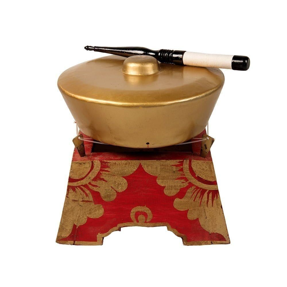 This is a product image of the Bonang - 1 Pan - Premium - Medium. It is a gold coloured pan with a gold boss on the top suspended by wire on a wooden frame. The frame is red with gold detail. There is a wooden beater that is wrapped in string at one end laying on the pan. The image has been taken from a head on position.