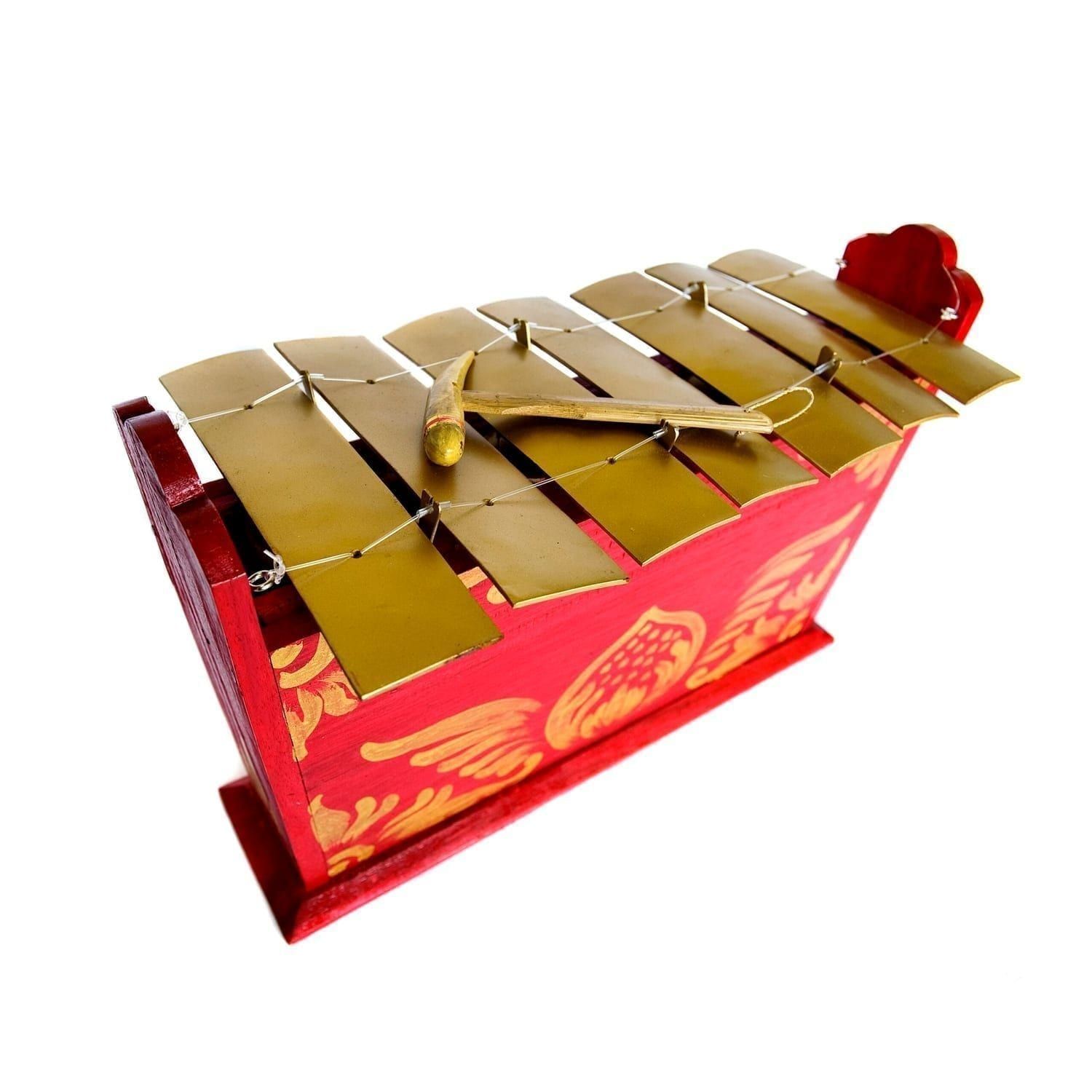 This is a product image of the Gamelan - Standard - Large - 7 key. It is red, with gold detail and has gold keys suspended from wire with a beater resting upon them. The image has been taken from above and the front with the instrument angled slightly to the right.