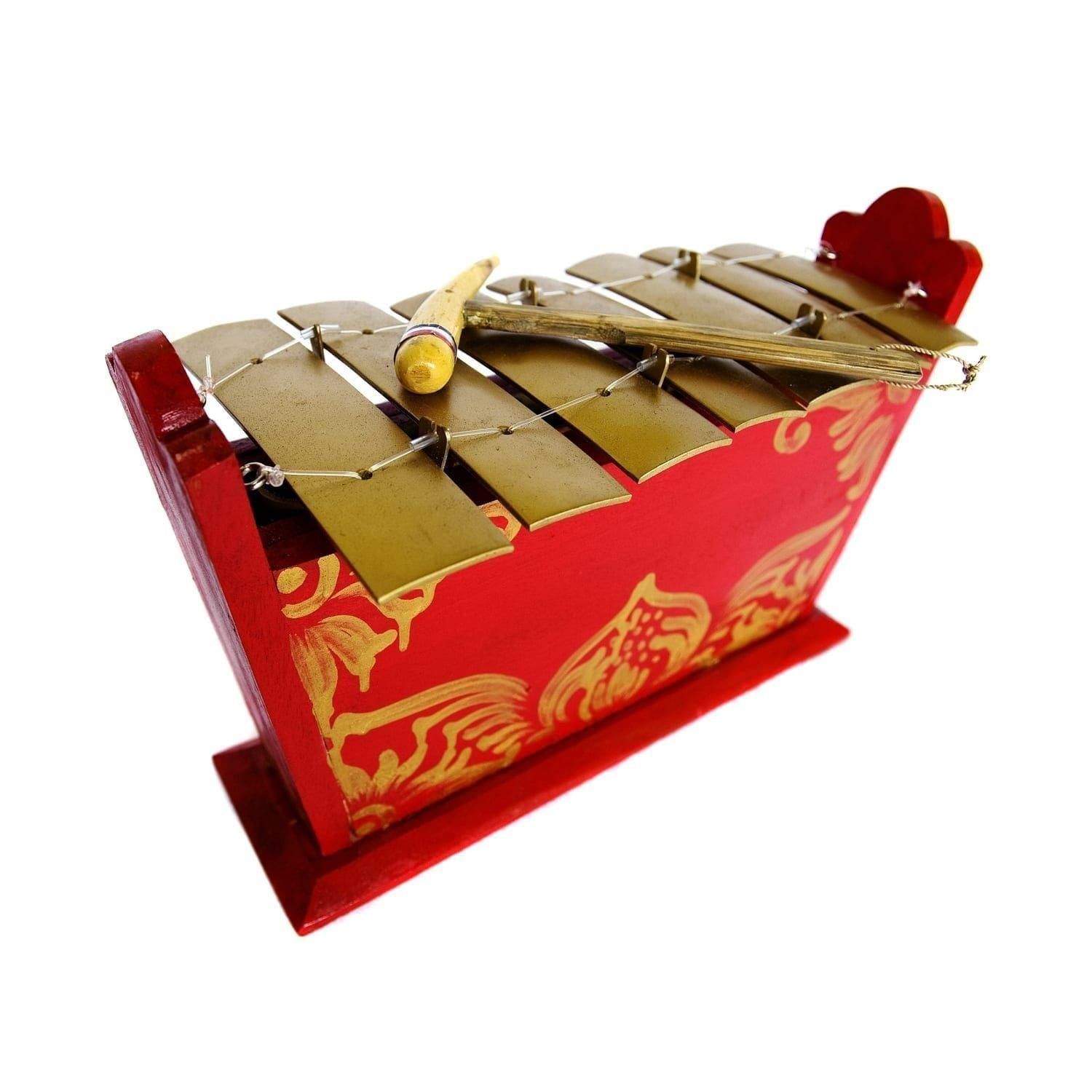 This is a product image of the Gamelan - Standard - Medium - 7 key. It is red, with gold detail and has gold keys suspended from wire with a beater resting upon them. The image has been taken from above and head on with the instrument facing to the right.