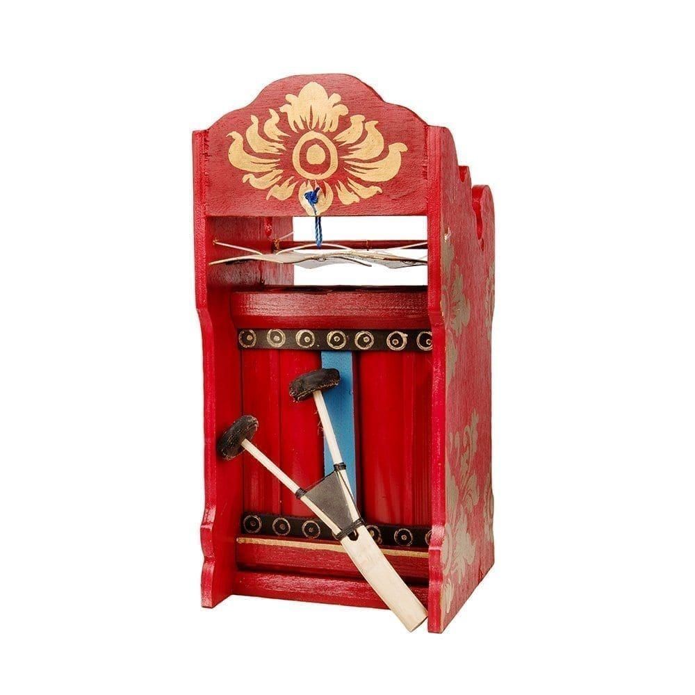 This is a product image of the Pulu- Budget. It is red with gold detail, and two gold bars suspended by wire. There is black, gold and blue detail on the instrument, and the two-headed beater is resting against the instrument in an upright fashion. The image has been take from head on with the instrument facing slightly to the left.