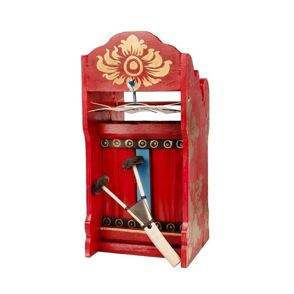 This is a product image of the Pulu- Budget. It is red with gold detail, and two gold bars suspended by wire. There is black, gold and blue detail on the instrument, and the two-headed beater is resting against the instrument in an upright fashion. The image has been take from head on with the instrument facing slightly to the left.