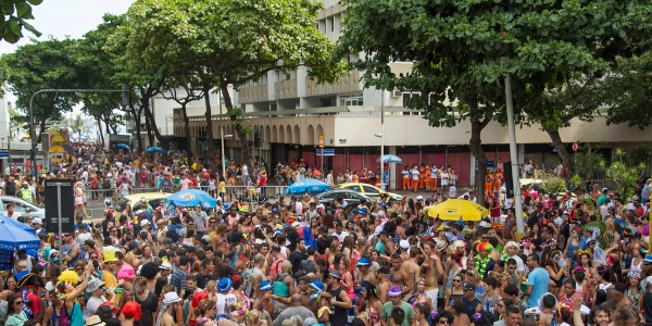 Crowded Rio street filled with Rio Carnival revellers in colourful costumes and hats, gathered under trees beside city buildings.